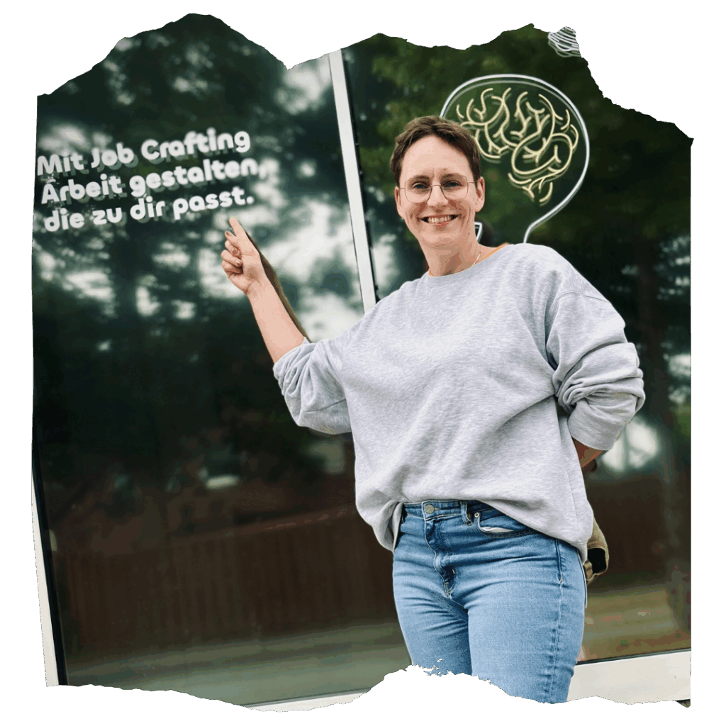 Antonia Ludwig steht vor einem Fenster mit der Aufschrift ‚Mit Job Crafting Arbeit gestalten, die zu dir passt.‘, zeigt darauf und lächelt in die Kamera.