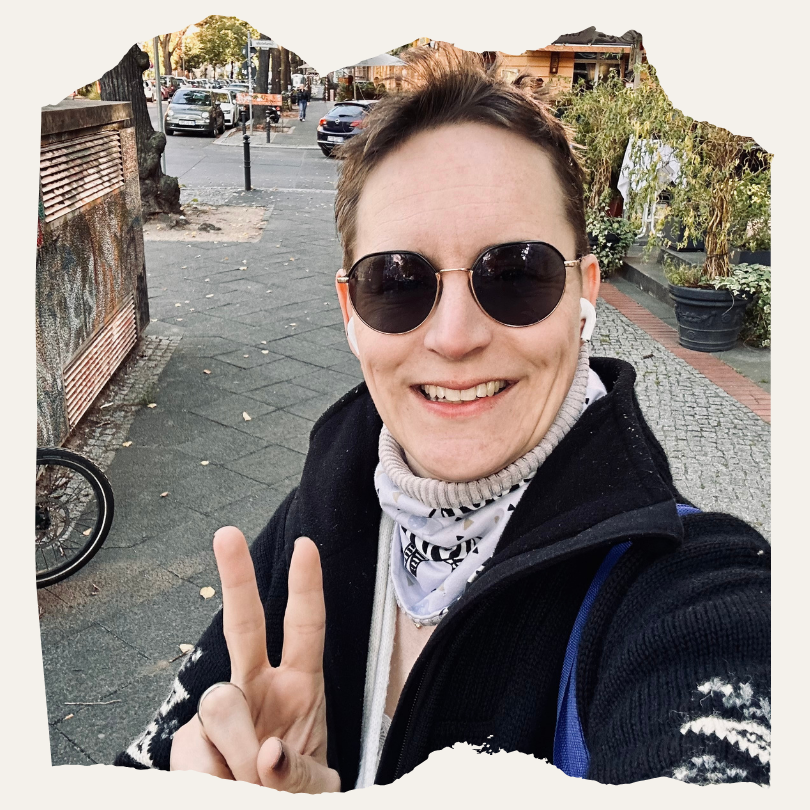 Eine Person macht ein Selfie auf einer Straße, trägt Sonnenbrille, Schal und Kopfhörer und zeigt ein Peace-Zeichen. Im Hintergrund stehen Autos und Bäume am Straßenrand.