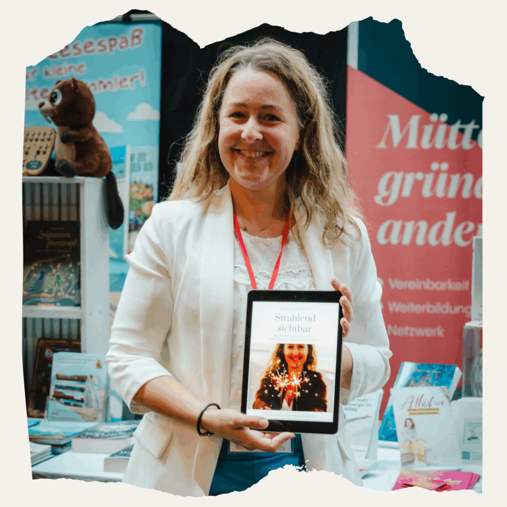 Eine Frau (Florence Chazarenc) steht an einem Messestand auf der BuchBerlin und hält ein Tablet mit dem Buchcover ‚Strahlend sichtbar‘ in den Händen. Im Hintergrund sind Bücher und ein Banner mit der Aufschrift ‚Mütter gründen anders‘ zu sehen.