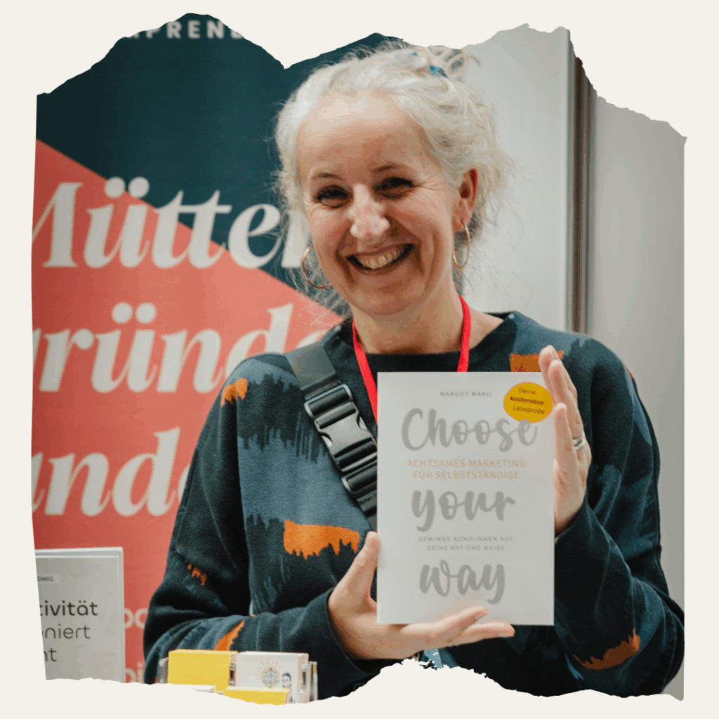 Eine lächelnde Frau (Margot Maric) steht am Mompreneurs-Stand auf der BuchBerlin und hält die Leseprobe ‚Choose your way – Achtsames Marketing für Selbstständige‘ in den Händen. Im Hintergrund ist ein Banner mit der Aufschrift ‚Mütter gründen anders‘ zu sehen.