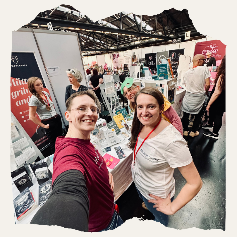 Zwei Personen stehen lächelnd am Mompreneurs-Stand auf der BuchBerlin und machen ein Selfie. Im Hintergrund sind Bücher, Banner und weitere Besucher:innen der Messe zu sehen.