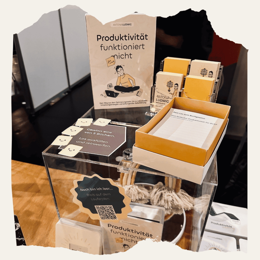 Messestand mit dem Buch ‚Produktivität funktioniert (für mich) nicht‘ von Antonia Ludwig. Daneben Gewinnspielkarten, Notizzettel und Buchflyer auf einem transparenten Aufsteller.