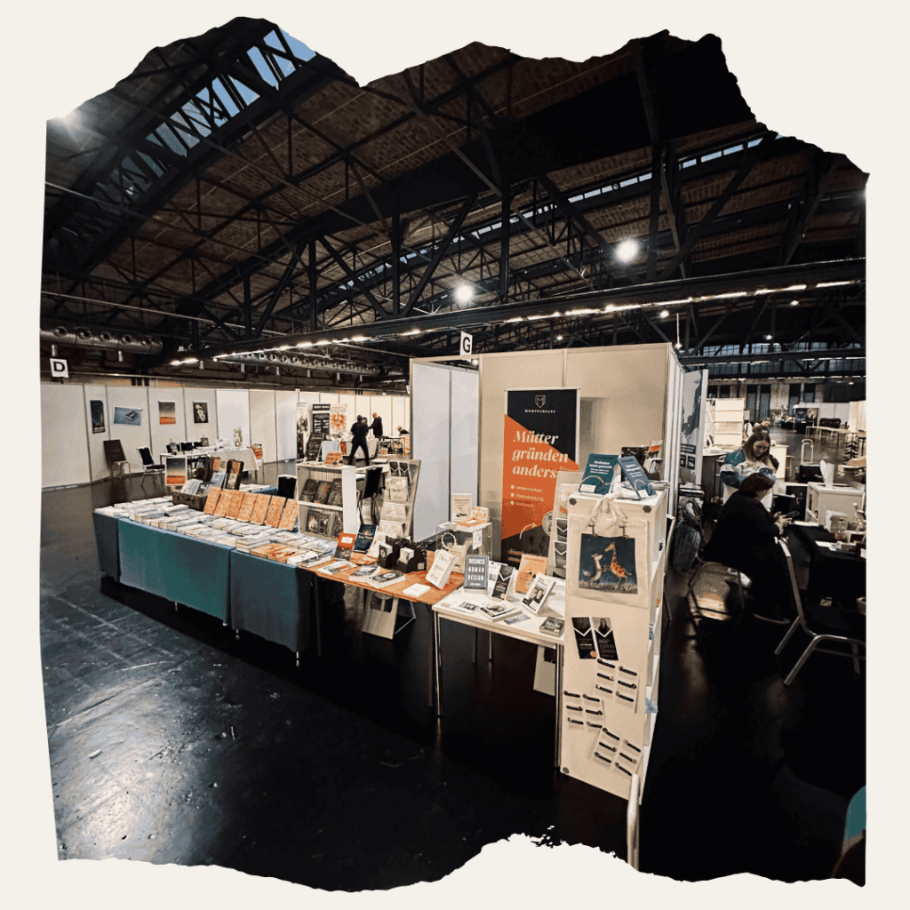 Blick in die Messehalle der BuchBerlin mit verschiedenen Verlagsständen. Im Vordergrund der Stand mit dem Banner ‚Mütter gründen anders‘ und zahlreichen ausgestellten Büchern und Taschen.