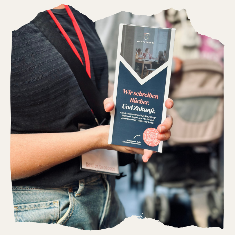 Eine Person hält einen Mompreneurs-Flyer mit der Aufschrift ‚Wir schreiben Bücher. Und Zukunft.‘ in der Hand. Im Hintergrund sind Besucher:innen der BuchBerlin zu sehen.