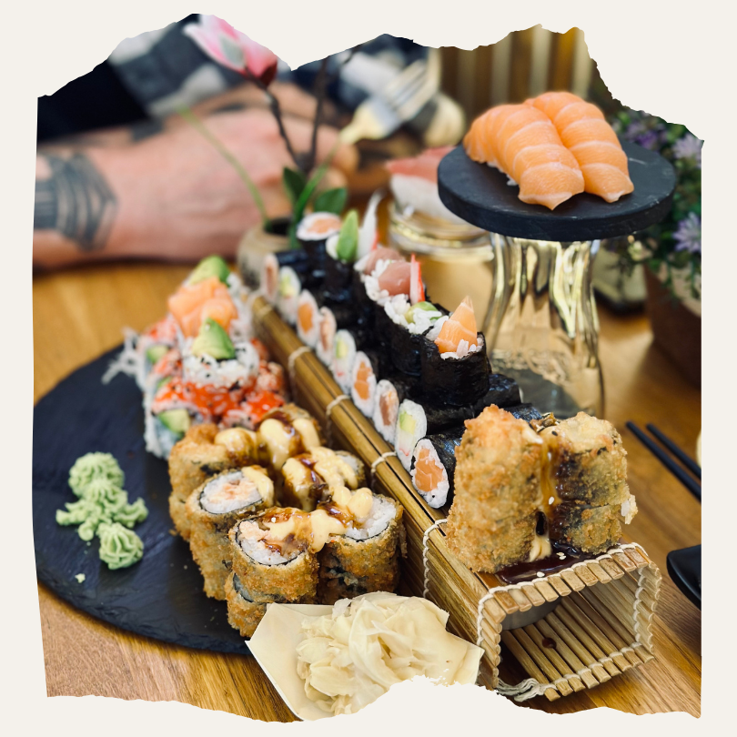 Eine Auswahl verschiedener Sushi-Rollen und Nigiri auf einem Holztisch, angerichtet auf Schieferplatten und Bambusmatten. Im Hintergrund ist eine Person mit tätowiertem Arm zu sehen.